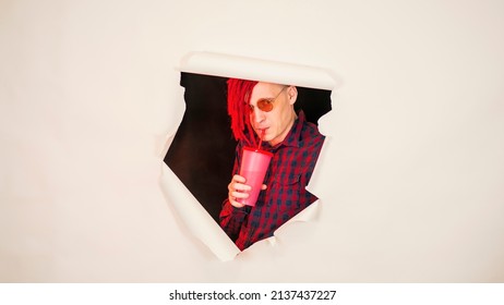 Young man in red plaid shirt drinking through soft drink straw in hole of white background. Bright guy enjoying nonalcoholic beverage - Shutterstock ID 2137437227