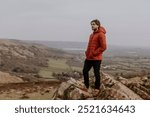 A young man in a red puffer jacket stands confidently on rocky terrain, enjoying the scenic view. Outdoor adventure with beauty of nature and spirit of exploration. Man hiking in Autumn nature.