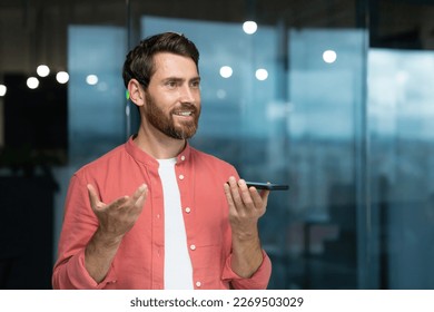 A young man in a red shirt sits in the office and talks on the phone through the loudspeaker, records the conversation on the recorder, voice message, looks away, smiles. - Powered by Shutterstock