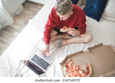 Young man in a red sweatshirt sits on a bed with a pizza box and a laptop, a view from above.Student sitting dmao on the bed eats a pizza and uses a laptop. Rest houses with internet, movie and pizza. - Powered by Shutterstock