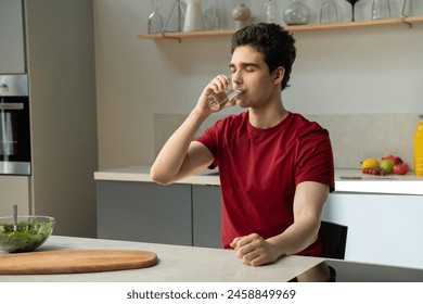 A young man in a red t-shirt stands in a well-lit, contemporary kitchen as he sips water from a clear glass. A healthy salad and various fruits are visible on the countertop, suggesting a focus on a - Powered by Shutterstock