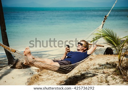 A young man relaxes in a hammock at the beach while checking messages on his smartphone. Toned image
