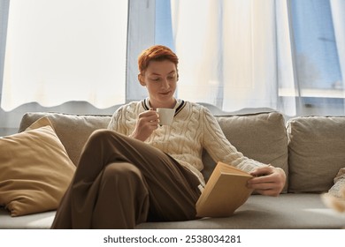 A young man relaxes on a couch, sipping tea while reading a book in a cozy setting. - Powered by Shutterstock