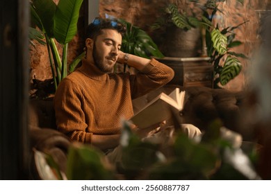 Young man relaxing with book in cozy cafe corner, surrounded by tropical plants and warm lighting - Powered by Shutterstock