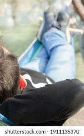 Young Man Relaxing Laying In Hammock At Home Outdoors In The Garden. Male Resting In Hammock Laying On Back, Arms Behind Head, Wearing Blue Jeans And Black Sweater