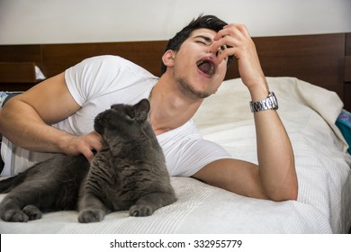 Young Man Relaxing On Bed Petting Grey Furry Cat And Sneezing From Allergies - Man Lying Down With Pet Cat And Having Allergic Reaction