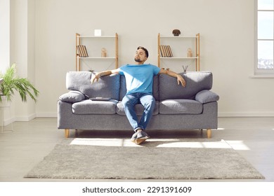 Joven relajándose en el sofá. El hombre disfruta su día libre o se detiene mientras trabaja. Hombre relajado sentado en un cómodo sofá con ordenador portátil, inclinado hacia atrás y disfrutando de un tranquilo tiempo solo en casa