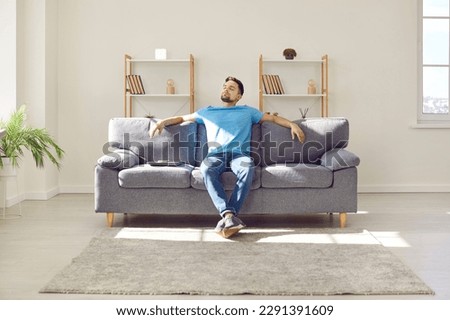 Young man relaxing on couch. Man enjoys his day off or takes pause while working. Relaxed man sitting on comfortable sofa with laptop, leaning back, and enjoying good quiet peaceful time alone at home