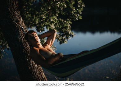 Young man relaxing on a hammock, enjoying a peaceful summer day in nature by the lake. A moment of tranquility and leisure. - Powered by Shutterstock