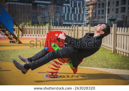 Young man reliving his childhood plying in a children's playground riding on a colorful red spring seat with a happy smile in an urban park