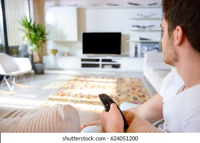 Young Man With Remote Controller Sitting Against Blank Scree Of TV In Living Room