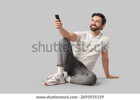 Young man with remote controller watching TV on light background