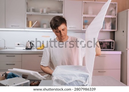 Young man removes protective packaging from recently delivered box of computer components on kitchen counter, preparing to assemble or upgrade system.