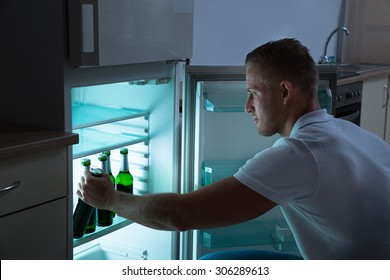 Young Man Removing Beer Bottle From Refrigerator At Night In Kitchen Room