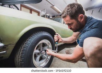 Young Man Removing Wheels Or Tires From An Old Vintage Car From The 60s Or 70s In His Home Garage. Tools Are Seen Around.