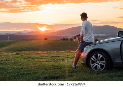 Young Man Resting During Long Solo Auto Trip Sitting On His Luxury Car Bonnet And Enjoying The Sunset Sky Colors. Traveling By Car Concept Image.