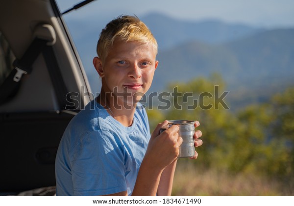 A\
young man is resting in nature with an off-road vehicle Drinks\
coffee from an iron mug. Resting in the trunk of a\
car
