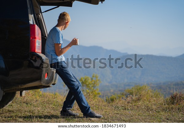 A
young man is resting in nature with an off-road vehicle Drinks
coffee from an iron mug. Resting in the trunk of a
car
