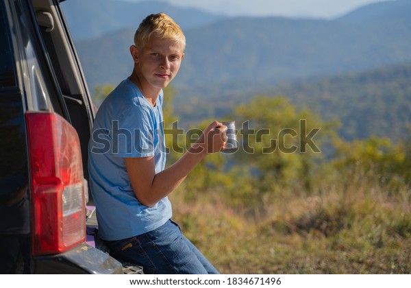 A\
young man is resting in nature with an off-road vehicle Drinks\
coffee from an iron mug. Resting in the trunk of a\
car