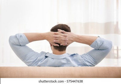 Young man resting on couch with hands behind his head in light room