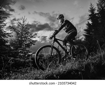 Young Man Riding Bicycle Downhill. Male Bicyclist In Sports Cycling Suit Cycling Down Grassy Hill At Night. Concept Of Sport, Biking And Active Leisure. Monochrome Image.