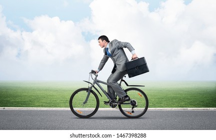 Young Man Riding Bicycle On Highway At Sunny Day. Businessman On Bike Hurry To Work. Corporate Employee Wearing Business Suit With Suitcase On Cycle Outdoor. Male Cyclist On Background Of Blue Sky