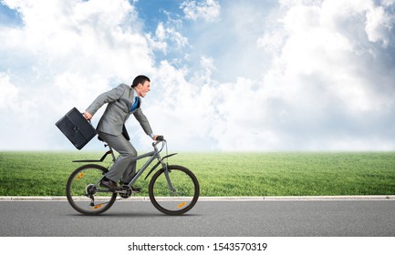 Young Man Riding Bicycle On Highway At Sunny Day. Businessman On Bike Hurry To Work. Corporate Employee Wearing Business Suit With Suitcase On Cycle Outdoor. Male Cyclist On Background Of Blue Sky