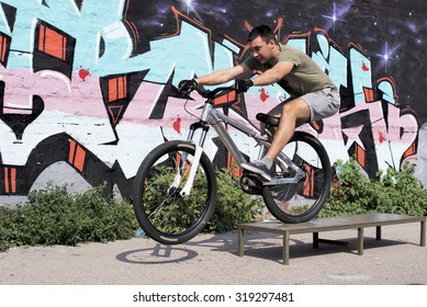 Young Man Riding A Bike, Doing A Trick In Front Of Graffiti Wall