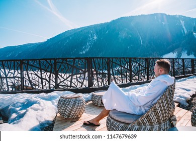 Young man in a robe chilling on a open terrace in the SPA with an amazing mountains view during winter time. Ski resort.