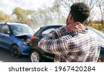 Young man rubbing neck in pain from whiplash injury standing by damaged car after traffic accident