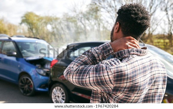 Young man rubbing neck in pain\
from whiplash injury standing by damaged car after traffic\
accident