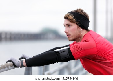 Young Man Runner Doing Running Warm-up Before Winter Run In City Outdoor. Athlete Wearing Smartwatch, Phone Armband For Music And Warm Sportswear For Cold Weather: Gloves, Headband, Long Underwear.