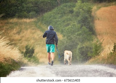 Young Man Running With His Dog (labrador Retriever) In Heavy Rain On The Rural Road.