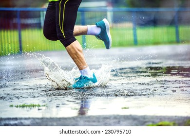 Young man running on asphalt sports field in rainy weather. Details of legs and sports shoes splashing in puddles.