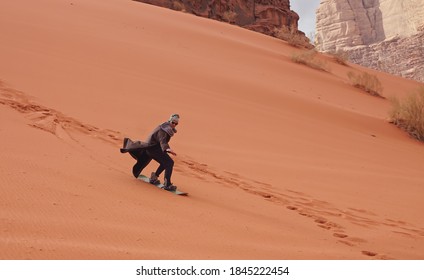 Young Man Sand Dune Surfing Wearing Bisht - Traditional Bedouin Coat. Sandsurfing Is One Of The Attractions In Wadi Rum Desert