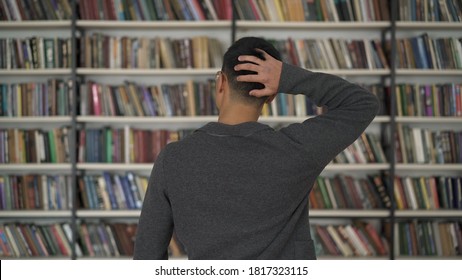 Young Man Scratching The Back Of His Head, Looking At The Bookshelves In Library. Choosing A Book, What Book To Buy, Hard Choice In A Big Library