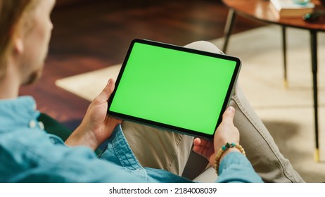 Young Man Scrolling and Tapping on Content on Tablet Computer with Green Screen Mock Up Display. Male Relaxing at Home, Reading Social Media Posts on Mobile Device. Close Up Over the Shoulder Photo. - Powered by Shutterstock