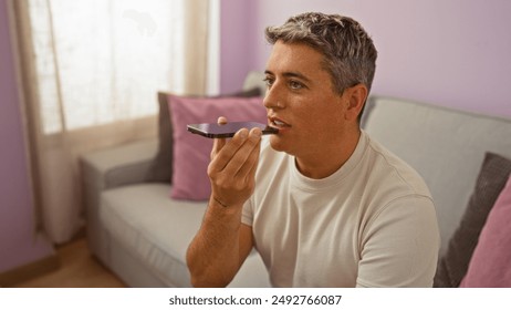 Young man seated in living room using a smartphone for voice command in a cozy indoor setting with pastel decor and natural light from a window - Powered by Shutterstock