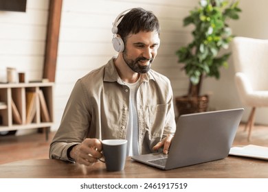 A young man is seated at a wooden table, engaging in remote work on his laptop. He wears headphones and holds a mug, suggesting a comfortable and casual work environment - Powered by Shutterstock