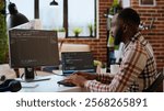 Young man seen programming software code on his laptop in a sleek home office. Workspace with dual monitors helps with software engineering, web development and coding productivity. Camera B.