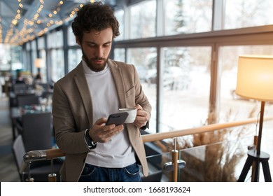 young man sending message to colleague. confident attractive young man holding smart phone and looking at it while standing in front of the big window in cafe, close up photo, free time, spare time - Shutterstock ID 1668193624