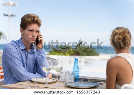 Young man with serious expression with glasses and piercings seated on a table talking with a mobile with a girl in front of him on a restaurant with seaviews