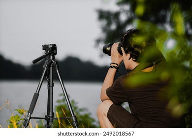 Young man setting up camera on the edge of the lake. Back view of male Setting up Camera with tripod