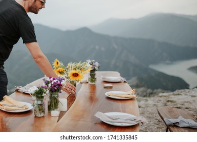 Young Man Setting Up Table For Picnic Outdoors. Mountain Top View And Canyon In Background