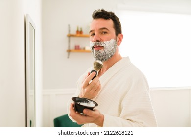 Young Man Shaving His Long Beard And Making Eye Contact. Bearded Hispanic Man Using Shaving Cream Foam