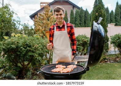 Young Man In A Shirt And An Apron Is Cooking Barbecue Meat On A Grill In His Home Garden. 