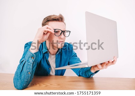 Young man in shirt and glasses he looks at the laptop in amazement and with a funny face sitting at the table on a white background. Funny face and amazement. Copy, empty space for text