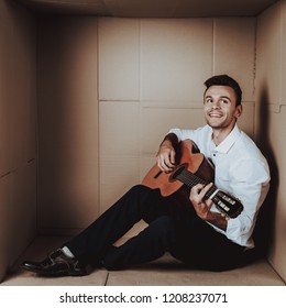 Young Man In Shirt Playing Guitar In Cardboard Box. Young Man In White Shirt. Life In Little Cardboard Box. Uncomfortable Life. Personal Spase Concepts. Young Introvert.