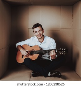 Young Man In Shirt Playing Guitar In Cardboard Box. Young Man In White Shirt. Life In Little Cardboard Box. Uncomfortable Life. Personal Spase Concepts. Young Introvert.