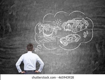 Young man in shirt and trousers is standing with back to viewer in front of blackboard with sketch of travel and car pictured on it. Concept of making dream come true. - Shutterstock ID 465943928